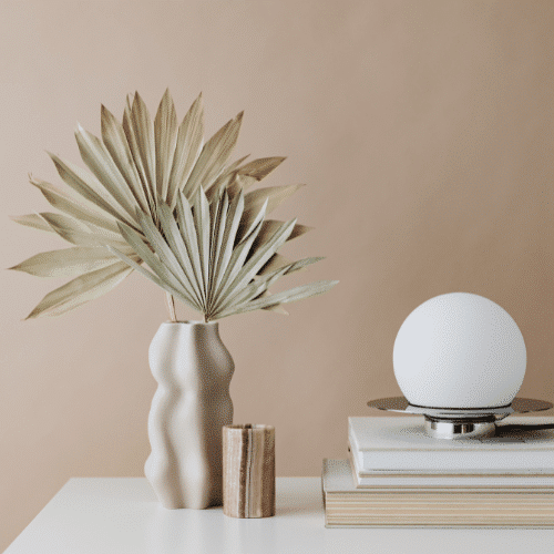 A dried palm leaves in a vase placed beside a round white modern table lamp on a light surface with beige coloured background.